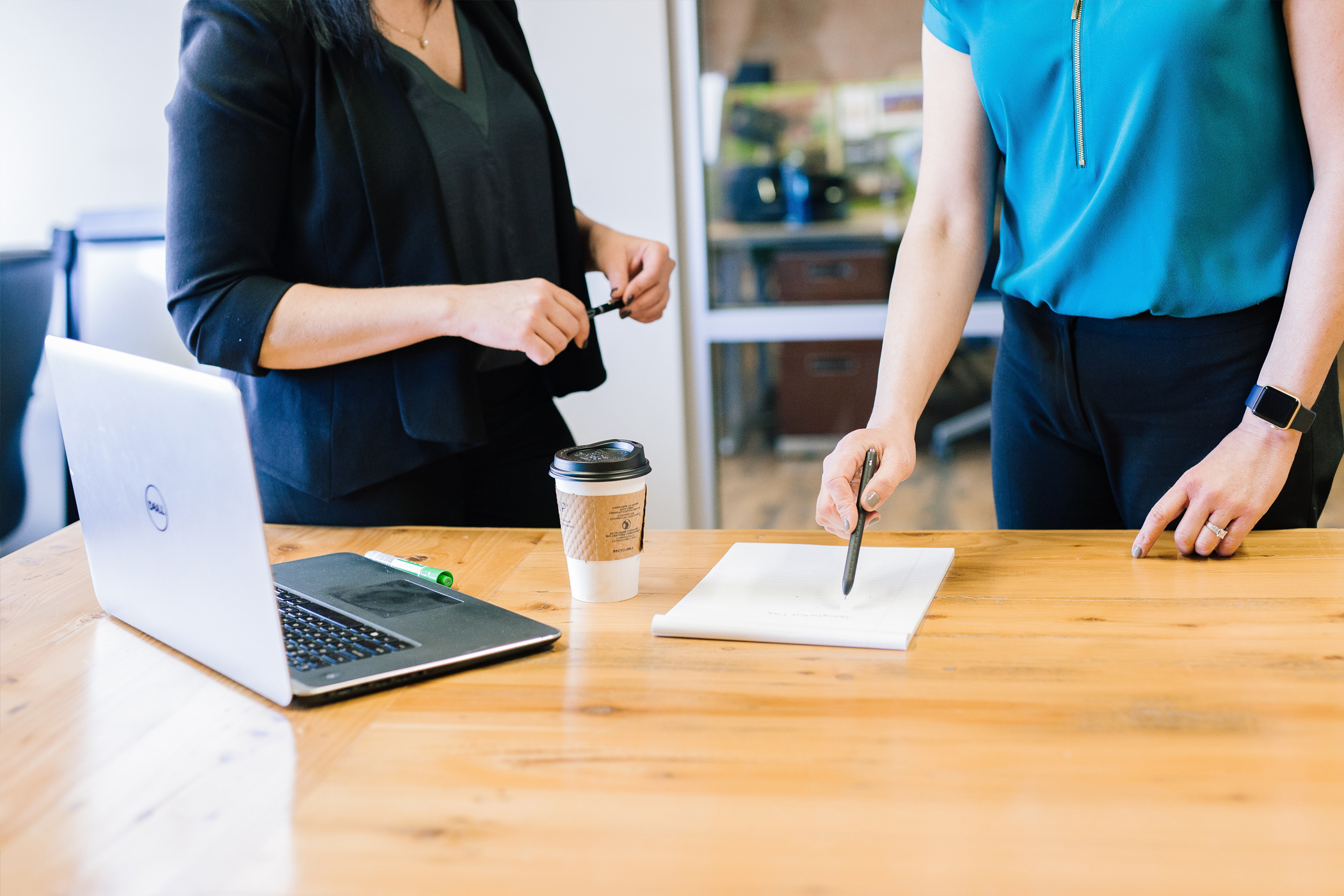 Two woman standing at a table with a laptop and notepad. One points with a pen to the notepad.